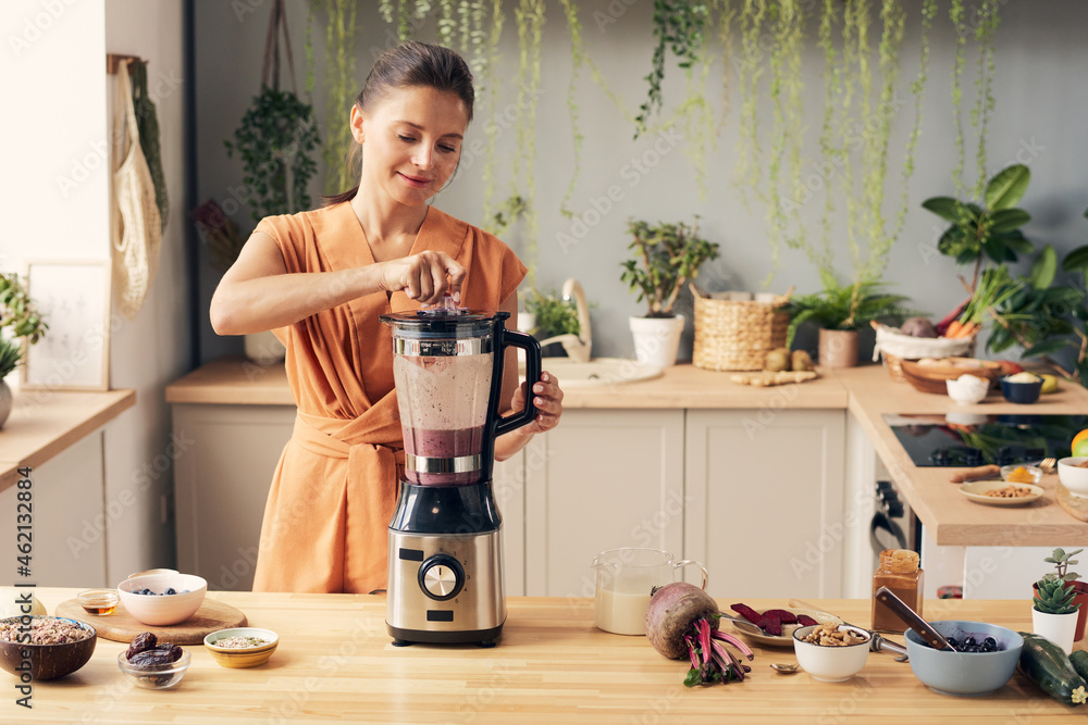 Young woman mixing ingredients of smoothie in electric blender while preparing healthy drink - obrazy, fototapety, plakaty 