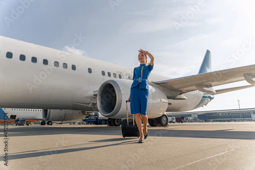 Smiling stewardess holding black suitcase and standing on runway with airplane in the background photo