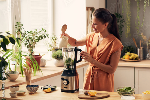 Happy young housewife pouring milk into electric blender while preparing homemade smoothie or yoghurt