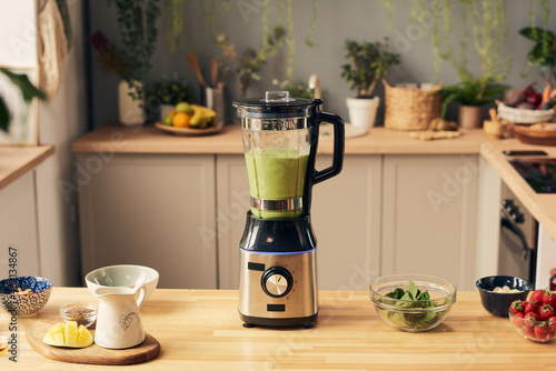 Electric blender with vegetable smoothie and bowls with its ingredients on kitchen table photo