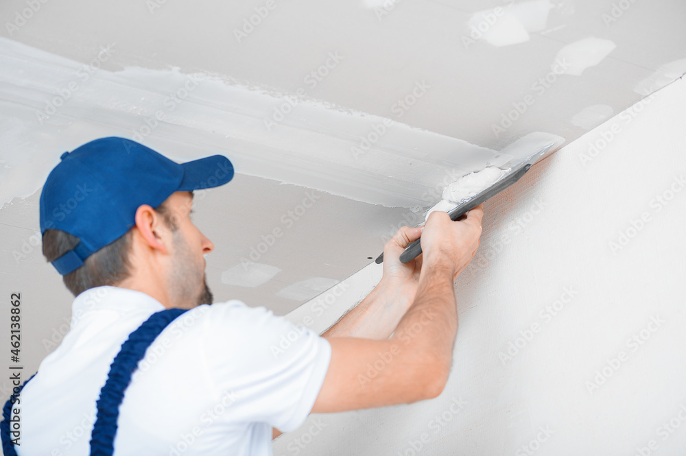 A master in uniform applies putty with a spatula to the reinforced joint of drywall on the ceiling. A master in uniform applies putty with a spatula to the reinforced joint of drywall on the ceiling.
