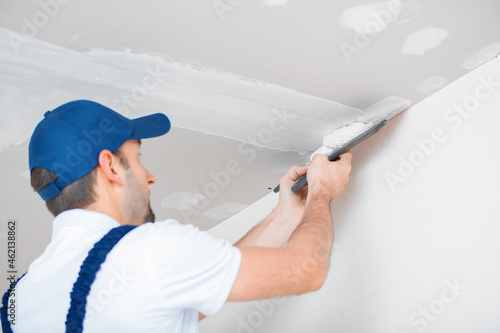 A master in uniform applies putty with a spatula to the reinforced joint of drywall on the ceiling.