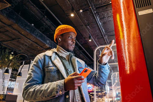 Obraz na plátně Smiling African-American man in warm denim jacket with wireless earphones uses s