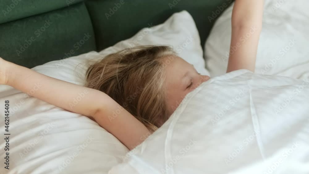 Cute little girl child wakes up in the morning in bed in the bedroom on an orthopedic mattress and pillow after a healthy sleep and does not want to wake up and hides under the covers. Cute little girl child wakes up in the morning in bed in the bedroom on an orthopedic mattress and pillow after a healthy sleep and does not want to wake up and hides under the covers.