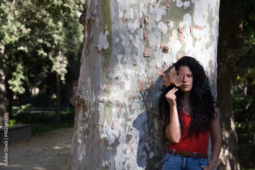 Fototapeta Naklejka Na Ścianę i Meble -  a beautiful, dark-haired, curly-haired adult woman with latin features is leaning against a giant tree. The woman covers her eye with a dry leaf that falls from the tree in autumn. Autumn concept