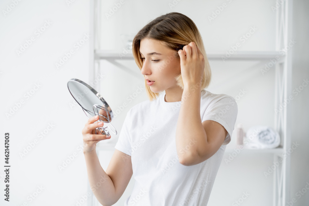 Hair care. Beauty treatment. Morning routine. Attractive young woman touching healthy soft locks looking in mirror in light bathroom.