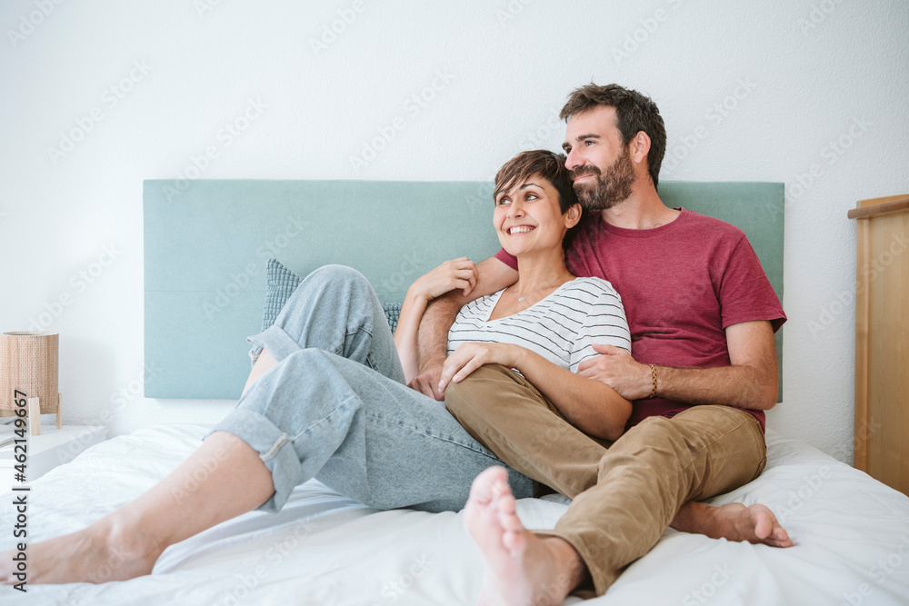 Happy mid adult couple looking away while sitting together on bed at home Happy mid adult couple looking away while sitting together on bed at home