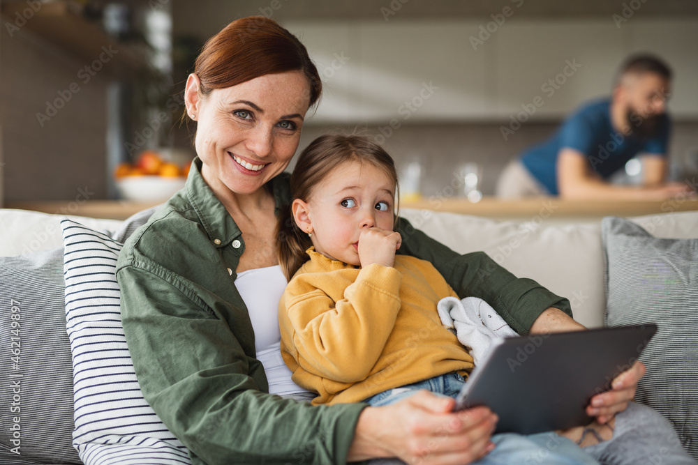 Mother with small daughter using tablet indoors at home, looking at camera, everyday life and home office with child concept. Mother with small daughter using tablet indoors at home, looking at camera, everyday life and home office with child concept.