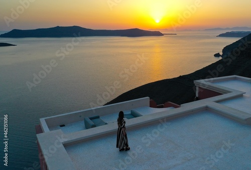 Sunset rooftop views in Santorini island, panoramic view of Aegean sea, popular touristic destination for honeymoon, woman in long dress watching sunset