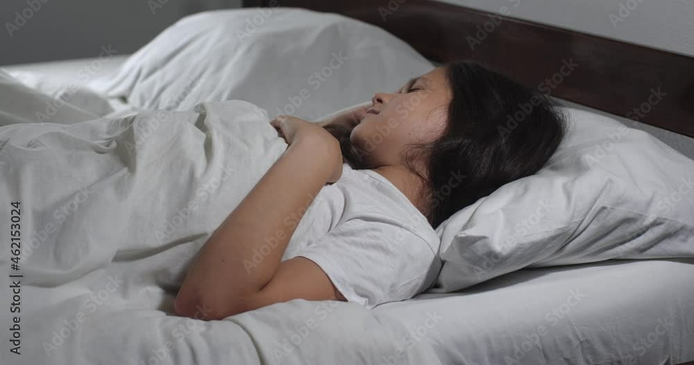 Close up of girl 9 years old with long brown hair lying in bed with white linens in light room suffering from headache. Teenager brunette suffers from pain migraine holding head with hands 4K Close up of girl 9 years old with long brown hair lying in bed with white linens in light room suffering from headache. Teenager brunette suffers from pain migraine holding head with hands 4K