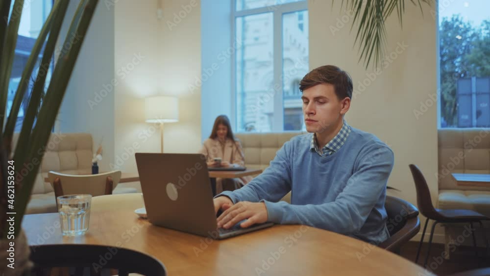 Young man wearing sweater sitting in cafe and typing on laptop, woman on blurred background using smartphone. Arc shot freelancer working remotely. Concept of self-employment Young man wearing sweater sitting in cafe and typing on laptop, woman on blurred background using smartphone. Arc shot freelancer working remotely. Concept of self-employment