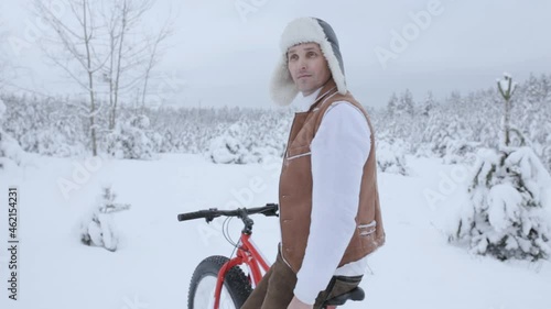 Male traveler with red fat bike standing in winter woods on snowy meadow and looking around while exploring natural environment 