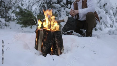 Crop male woodcutter warming hands near burning bonfire with orange flame in snowy meadow in winter woods 