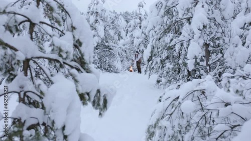 Traveling man in warm clothes warming hands over hot flam of burning campfire in snowy winter forest among evergreen trees 