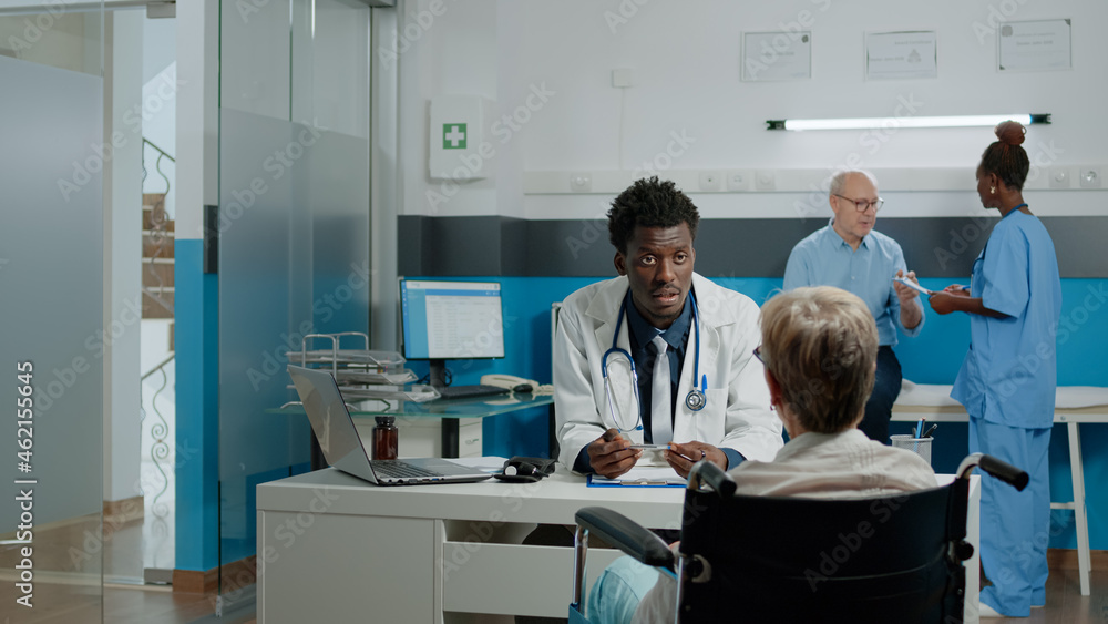 Elder patient with disability having checkup appointment with young doctor at medical clinic. Invalid old woman in wheelchair sitting at desk with medic while talking about healthcare problems Elder patient with disability having checkup appointment with young doctor at medical clinic. Invalid old woman in wheelchair sitting at desk with medic while talking about healthcare problems