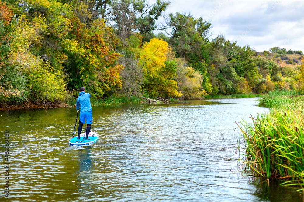 A man on a SUP swims along the river in autumn in cloudy weather