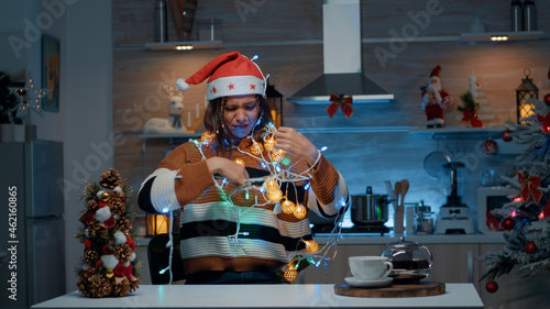 Smiling woman with santa hat tangled in christmas lights string while doing decor preparations in kitchen. Young person having fun indoors with festive decorations and ornaments
