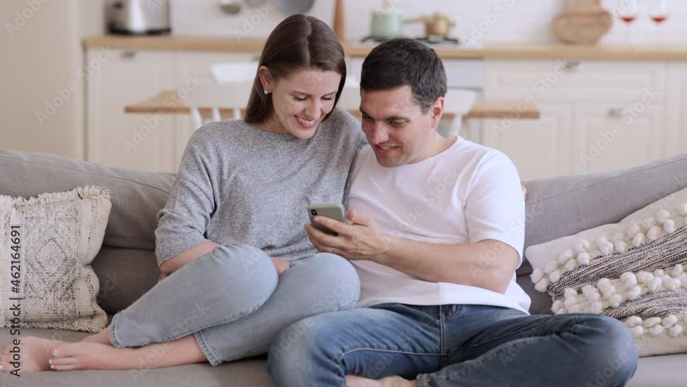 Young woman, man using smartphone smiling while sitting on sofa in home room spbi. Front view of beautiful woman, man hold phone in his hand and look at screen, talk with happy smiles and sit on couch Young woman, man using smartphone smiling while sitting on sofa in home room spbi. Front view of beautiful woman, man hold phone in his hand and look at screen, talk with happy smiles and sit on couch