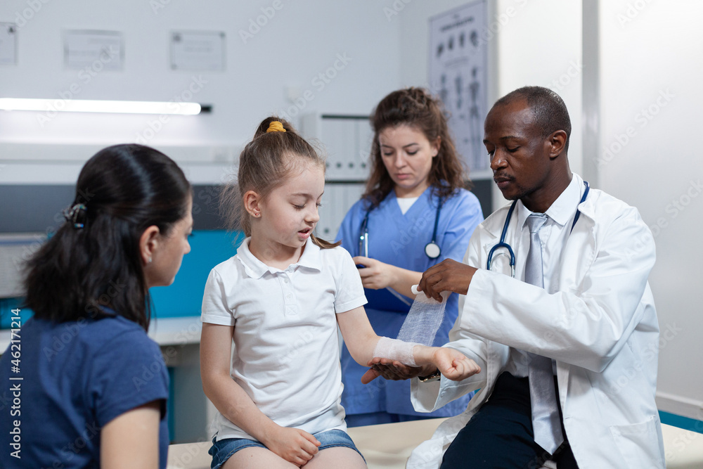 African american pediatrician doctor bandage broken arm of young child during medical appointment in hospital office. Girl patient with fractured hand having physiotherapy. Trauma recovery African american pediatrician doctor bandage broken arm of young child during medical appointment in hospital office. Girl patient with fractured hand having physiotherapy. Trauma recovery
