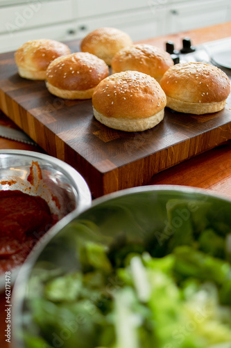 Technique for making burgers at home. Baked buns with sesame seeds in the kitchen on a wooden board. Lettuce leaves and red sauce in blur.