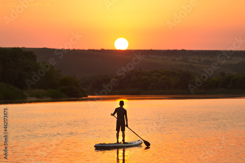 A man on a SUP on the river in the summer admires the sunset. Silhouette of a man on a sup on the background of the river in the rays of the sunset