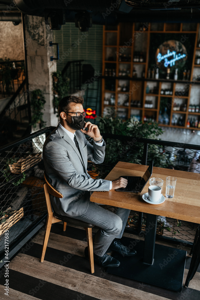 A businessman with a protective mask on his face is sitting in a cafe and talking on the phone while using laptop. Coronavirus, Covid-19 concept.