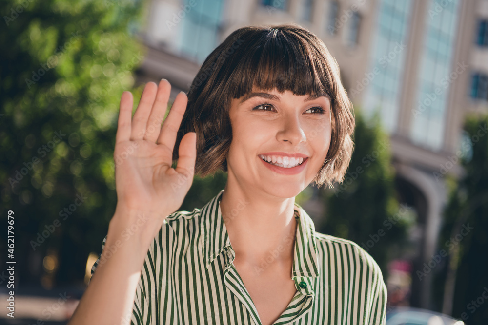 Photo of friendly positive cute lady raise hand wave palm beaming smile wear striped shirt urban city outside