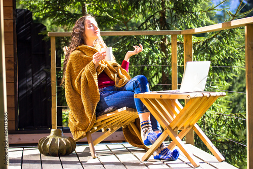 Pretty girl sitting on balcony terrace wrapped in yellow plaid holding cup of tea coffee, working remotely at laptop. Young brunette woman shopping online. House in pine forest at sunny autumn day.