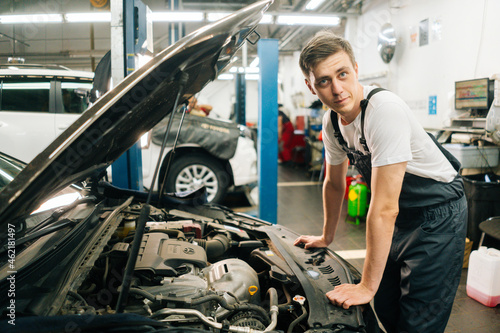 Side view of confident handsome professional male car mechanic in blue uniform standing in front of open hood, inspecting engine of car coming in for repair or maintenance, looking at camera.