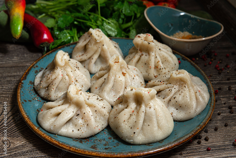 Georgian-style khinkali with meat on a handmade ceramic plate. Sprinkled with ground pepper. Leaves of parsley and coriander in the background. On a dark grey wooden kitchen table. Georgian-style khinkali with meat on a handmade ceramic plate. Sprinkled with ground pepper. Leaves of parsley and coriander in the background. On a dark grey wooden kitchen table.