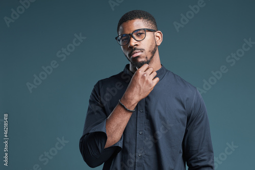 Waist up portrait of confident African-American man wearing glasses looking at camera while standing against deep blue background, copy space
