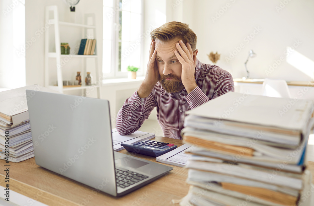 Tired exhausted male office worker working all day on financial report counting figures. Man holds his head sitting in front of laptop between large number of folders with documents. Overwork concept. Tired exhausted male office worker working all day on financial report counting figures. Man holds his head sitting in front of laptop between large number of folders with documents. Overwork concept.