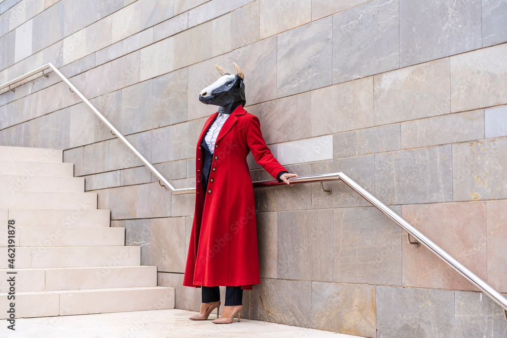 Woman wearing red overcoat and bull mask holding railing while standing near staircase Woman wearing red overcoat and bull mask holding railing while standing near staircase