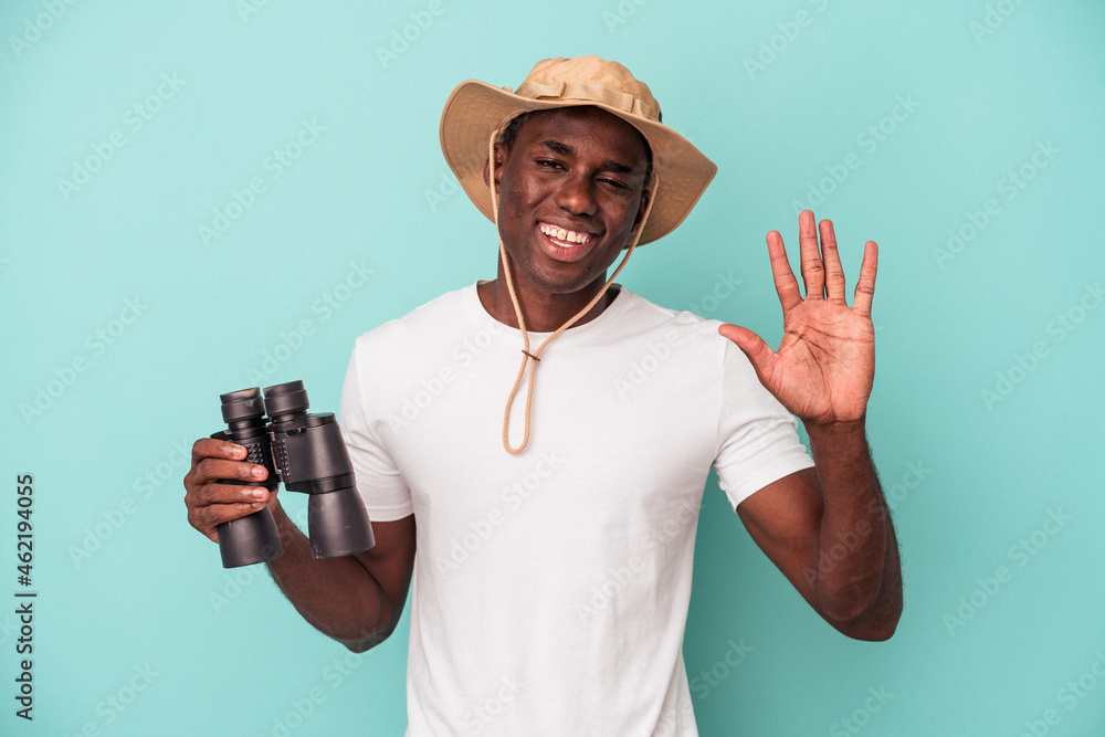 Young African American man holding binoculars isolated on blue background smiling cheerful showing number five with fingers. Young African American man holding binoculars isolated on blue background smiling cheerful showing number five with fingers.