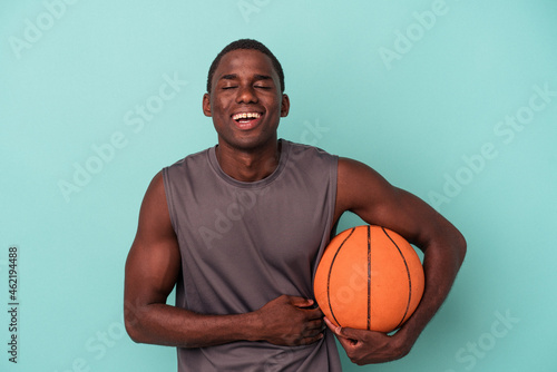 Young African American man playing basketball isolated on blue background laughing and having fun.