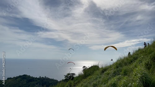 BASE jumping. Mountain, covered by green tropical forest. Sunlight. Beautiful sky with pattern of white clouds. Sea. Shine of sun on the surface of the water. People fly over the ocean by parachutes photo