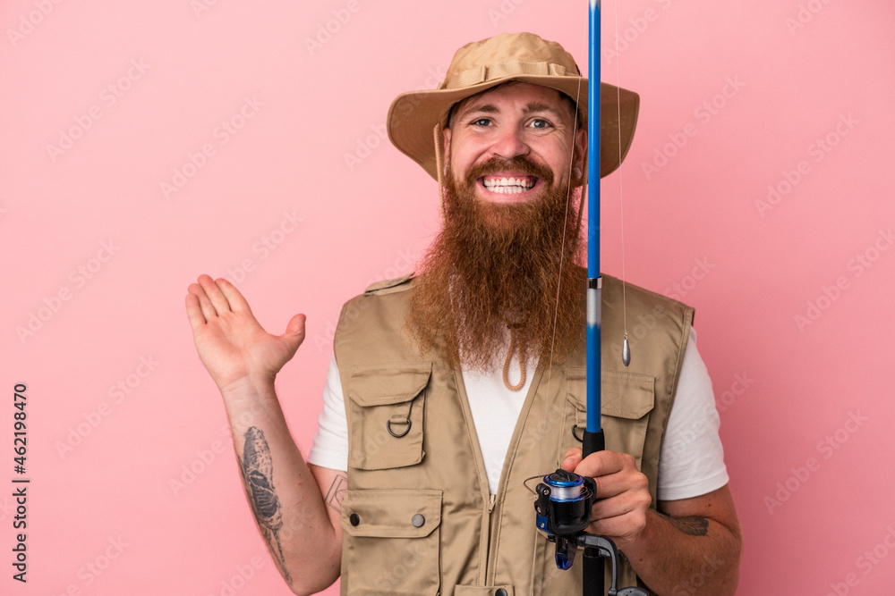 Young caucasian ginger fisherman with long beard holding a rod isolated on pink background showing a copy space on a palm and holding another hand on waist. Young caucasian ginger fisherman with long beard holding a rod isolated on pink background showing a copy space on a palm and holding another hand on waist.