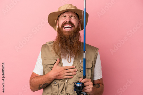 Young caucasian ginger fisherman with long beard holding a rod isolated on pink background laughs out loudly keeping hand on chest. photo