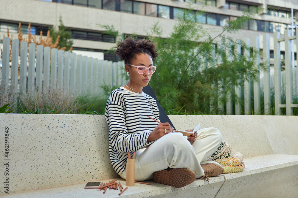 Serious creative female designer draws sketches with colorful pencils in notepad sits crossed legs dressed in stylish clothes poses against urban background. Afro American girl creats picture
