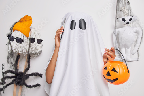 Indoor shot of spooky faceless ghost makes phone call holds carved orange pumpkin has scary image poses against white background with creepy toys around. Happy Halloween concept. Holiday mystery