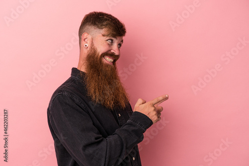 Young caucasian ginger man with long beard isolated on pink background points with thumb finger away  laughing and carefree.
