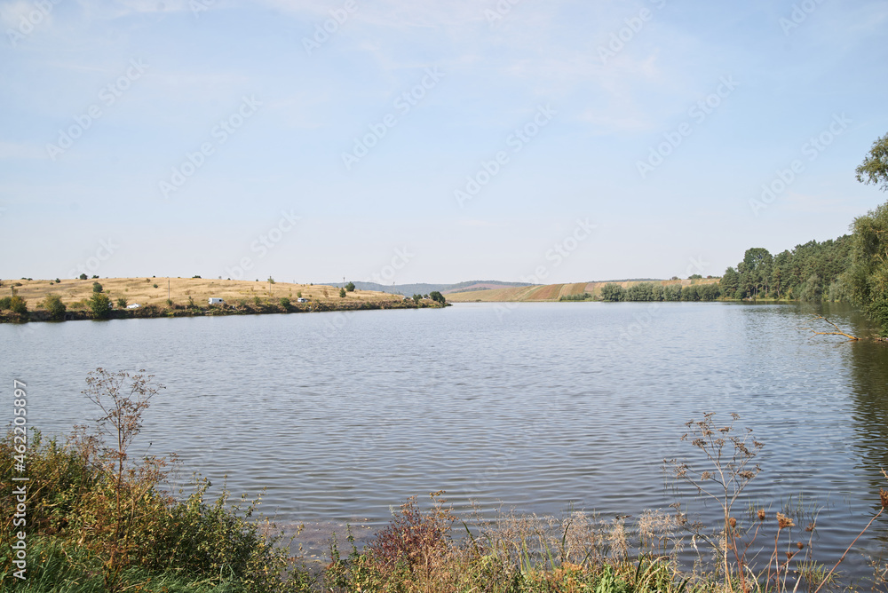 Amazing lake landscape with dry grass and green trees in autumn.Light blue sky over pond. Nature protection concept. Rural scene in countryside. Fish farming basin in the village.