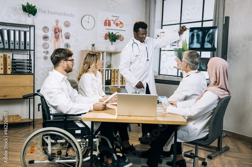 Group of multiracial medical specialists sitting together at office desk and listening african colleague that pointing on x ray scan. Competent doctors examining results of patient analysis.