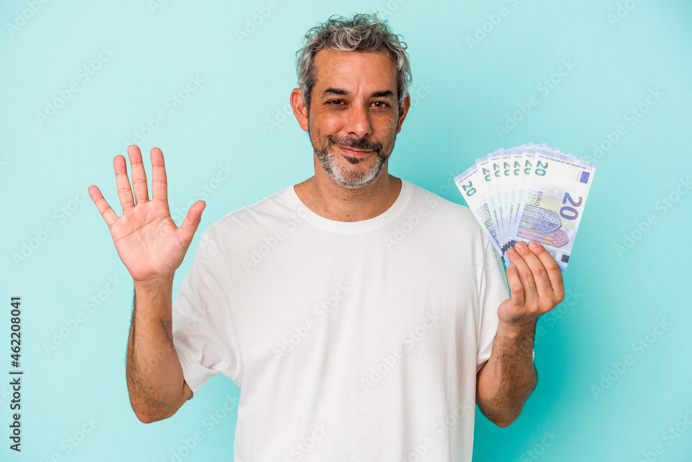 Middle age caucasian man holding bills isolated on blue background smiling cheerful showing number five with fingers. Middle age caucasian man holding bills isolated on blue background smiling cheerful showing number five with fingers.