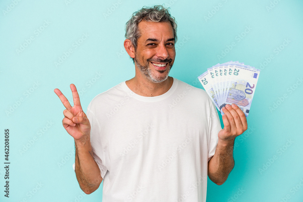 Middle age caucasian man holding bills isolated on blue background joyful and carefree showing a peace symbol with fingers. Middle age caucasian man holding bills isolated on blue background joyful and carefree showing a peace symbol with fingers.