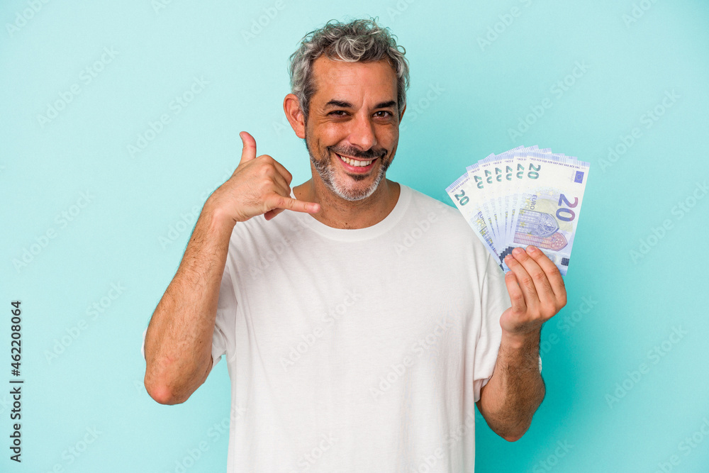 Middle age caucasian man holding bills isolated on blue background showing a mobile phone call gesture with fingers. Middle age caucasian man holding bills isolated on blue background showing a mobile phone call gesture with fingers.