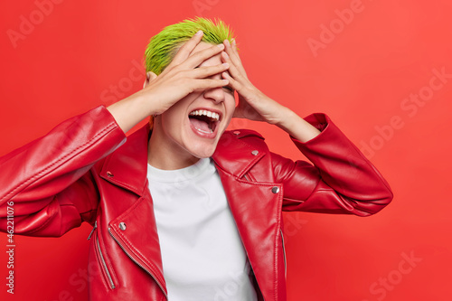 Slika na platnu Indoor shot of extraordinary hipster girl laughs happily tries to hide her face has green hairstyle dressed in casual leather jacket isolated over vivid red wall