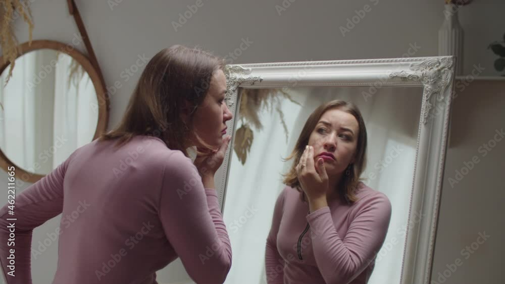 Portrait of beautiful displeased adult woman looking at herself reflection in mirror, concerned about wrinkles and skin aging, expressing worry and disappointment about her appearance at home. Portrait of beautiful displeased adult woman looking at herself reflection in mirror, concerned about wrinkles and skin aging, expressing worry and disappointment about her appearance at home.