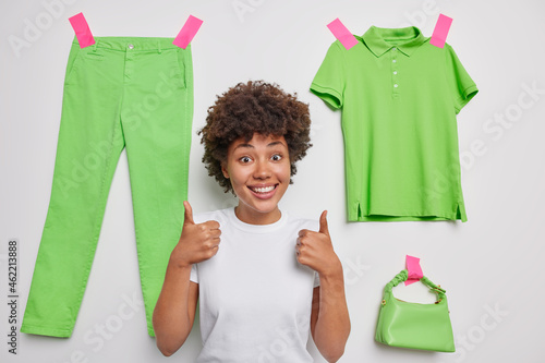 Positive young African American woman keeps thums up shows like gesture being in good mood poses against white background with plastered green trousers t shirt and bag recommends to buy items