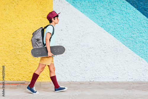 Boy with backpack and skateboard walking by multi colored wall photo